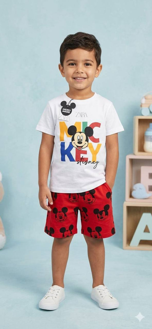 Child wearing a white t-shirt with colorful text and red shorts with black patterns, standing against a light blue background.